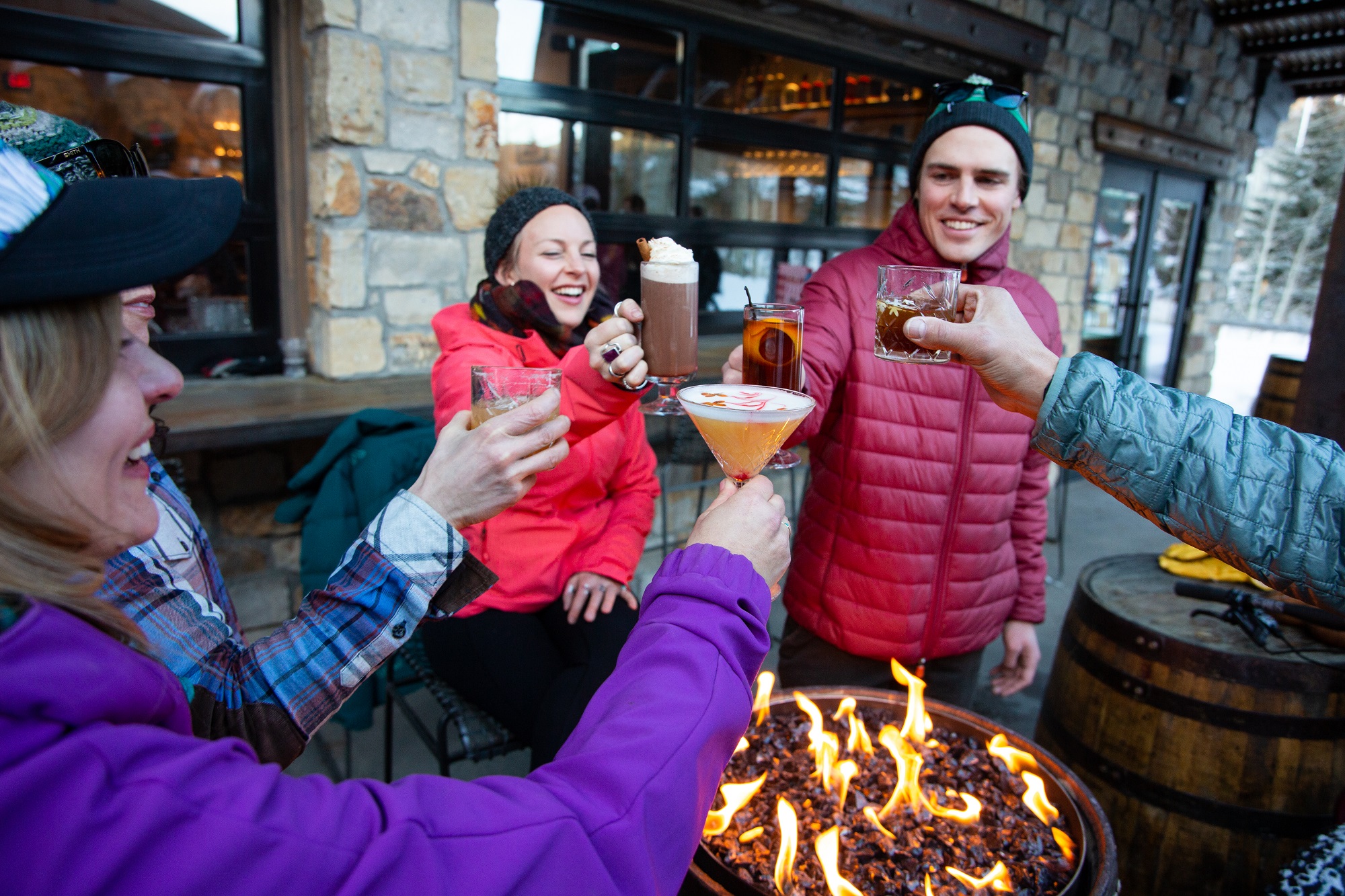 Group lifts cocktails up in celebration around an outdoor firepit.