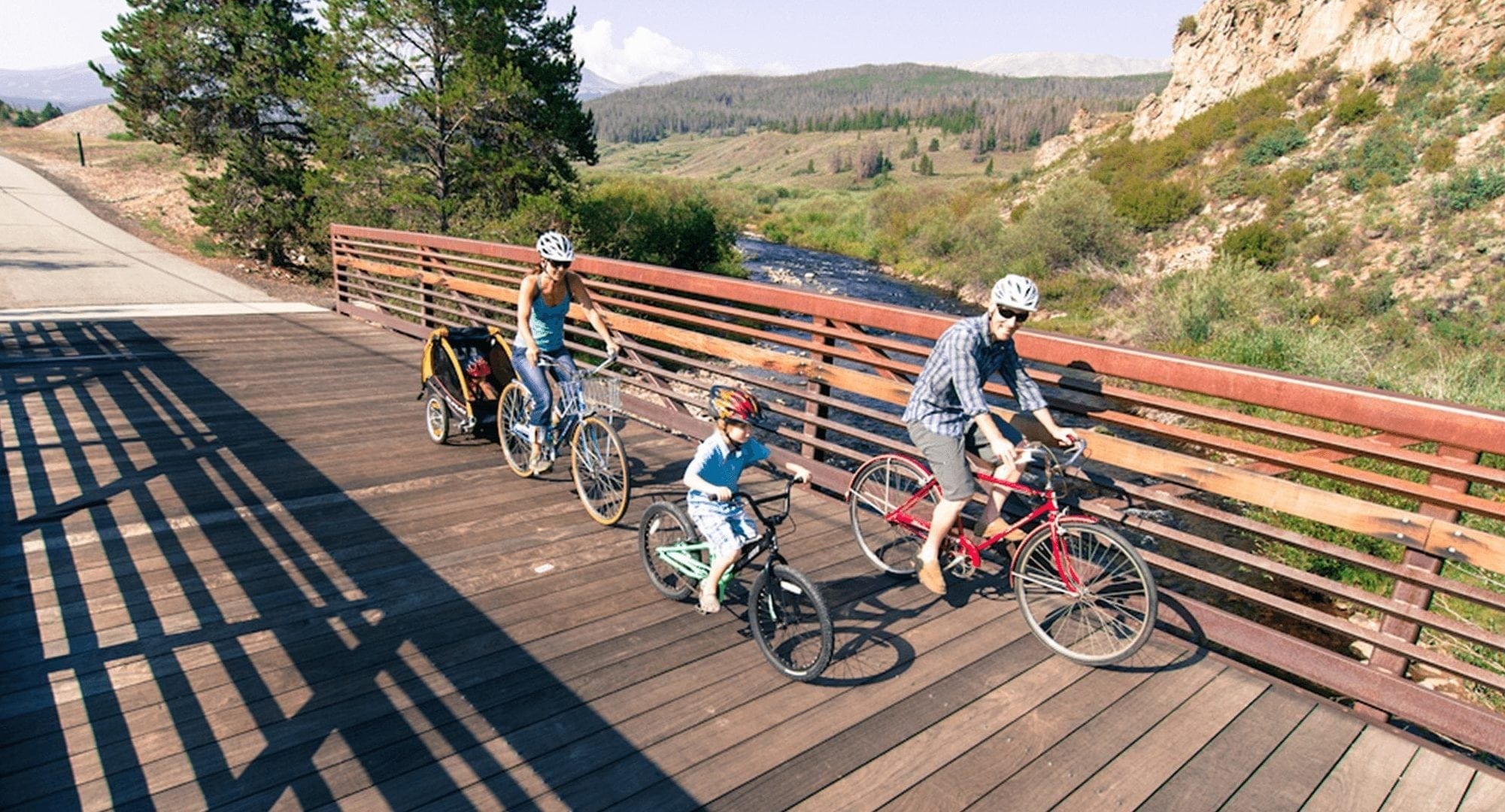 Family biking across a wooden bridge on a biking trail in Breckenridge, the Blue River Rec Path.