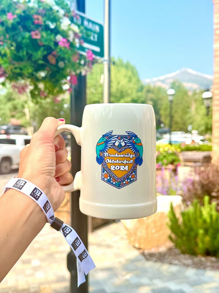 Holding the new Oktoberfest 2024 mug which features two bright blue hummingbirds and an orange heart. Breckenridge's Peak 8 is in the background along with stunning potted flowers.